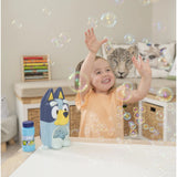 A smiling toddler with raised hands plays with floating bubbles in a bright nursery. A blue and yellow dog-shaped bubble machine and a small bubble bottle sit on a white table in the foreground, with a playroom bench and animal-print pillows in the background.