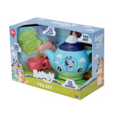 Bluey tea set in its retail packaging, featuring a light blue teapot with a dark blue lid, a pink cup, green plates, and other accessories visible through a clear plastic window.