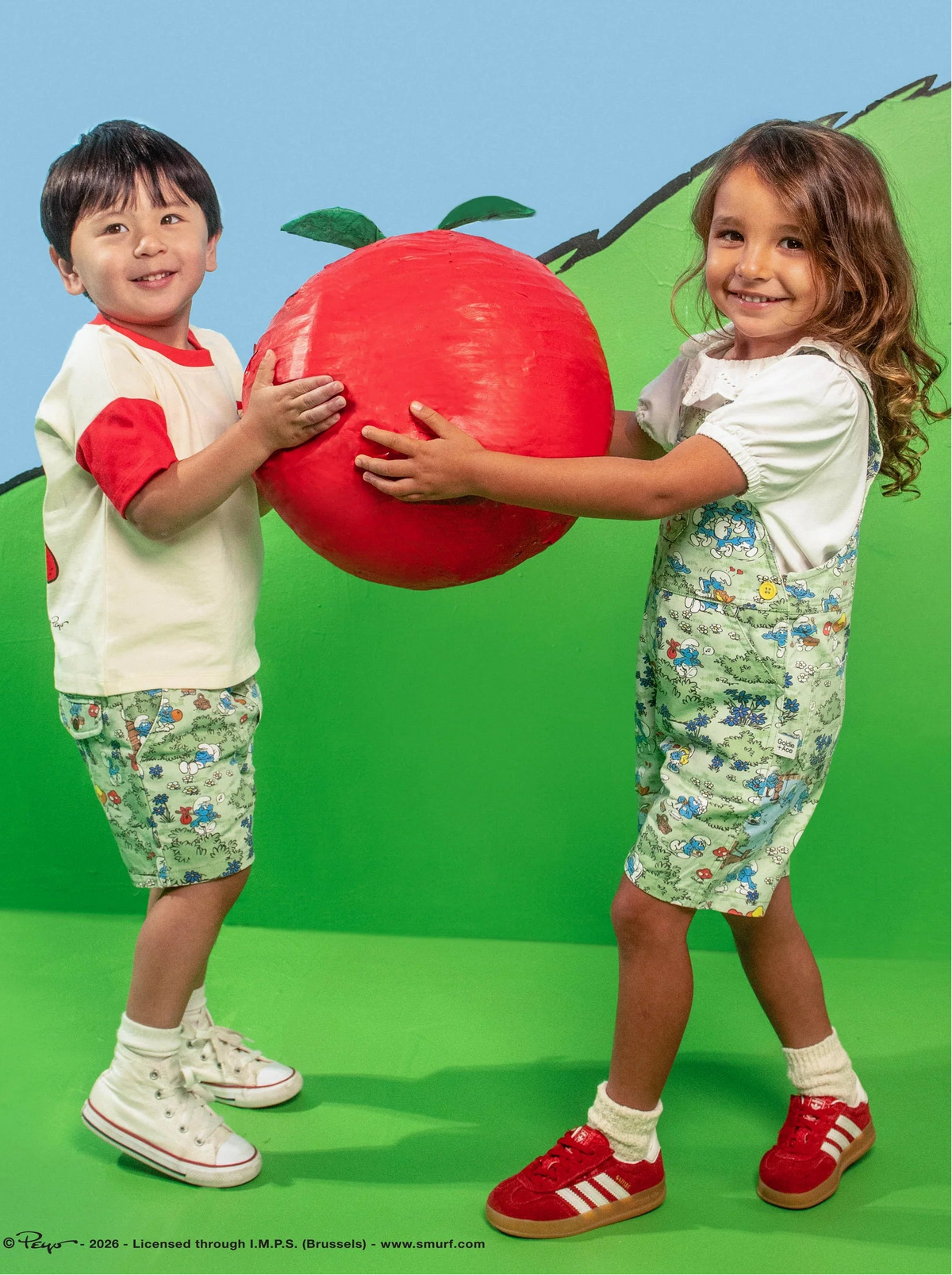 Two children wearing smurf printed overalls and shorts, holding a large red ball against a green and blue background