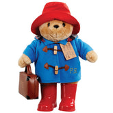 A large plush toy of Paddington Bear wearing his classic blue duffle coat, red hat, and wellington boots with the initials PB embroidered on them. He is also carrying a brown suitcase and has a tag around his neck that reads 'Please look after this bear, THANK YOU'.