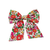 A fabric hair bow featuring a colorful retro floral pattern with red, pink, yellow, and green flowers on a white background, photographed against a neutral light gray backdrop.