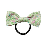A small bow tie made of light green fabric with a pink and green floral print, attached to a black elastic hair tie, shown against a plain light gray background.