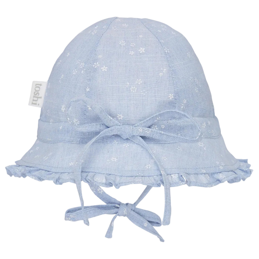 A light blue fabric baby sun hat with a white floral pattern, featuring a wide brim with ruffled trim, a matching bow on the front, and chin ties. A small fabric tag on the side reads 'loohi'.