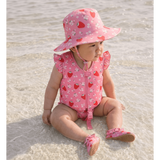 A baby sitting in shallow water on a beach, wearing a pink strawberry and white flower print sun hat, a matching pink ruffle-shoulder one-piece swimsuit with a front zipper, and pink slip-on beach shoes.