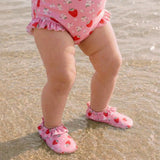 A close-up of a child standing in shallow water at the beach, wearing a pink one-piece swimsuit with a strawberry and white flower print, along with matching pink slip-on wet beach shoes.
