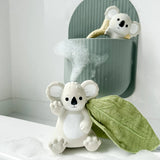 White koala toy with a green leaf on a white surface with another koala teether in a green tub on the wall of the bathroom