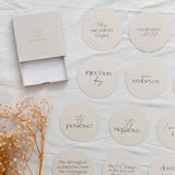 Round milestone plaques with text on a light surface with dried flowers besie teh box they come in.