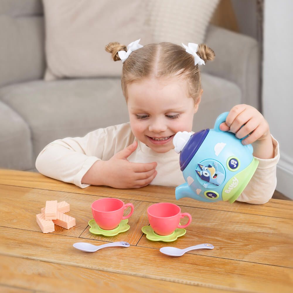 A young child with pigtails is smiling and pouring from a blue toy teapot with a cartoon dog design. In front of the child on a wooden table are two pink toy cups with green saucers, two white spoons, and two pink blocks.