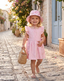 A young girl walking barefoot on a cobblestone street wearing a light pink dress with ruffled sleeves, a matching pink sun hat, and carrying a small woven straw bag.