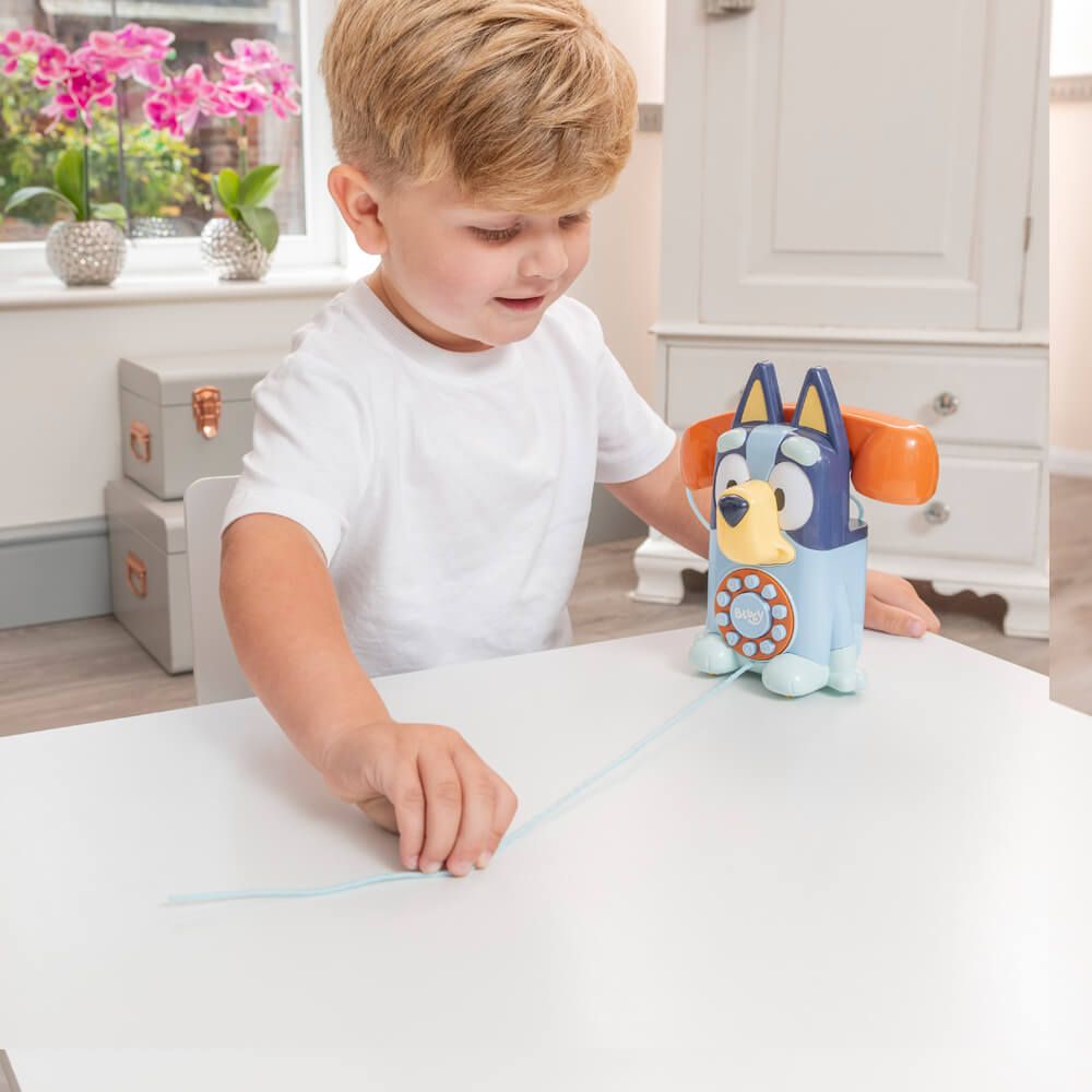 A young boy with blond hair wearing a white t-shirt is sitting at a white table, interacting with a Bluey-themed toy phone. The toy features a blue and yellow dog character with orange ear-like antennas and a light blue cord.