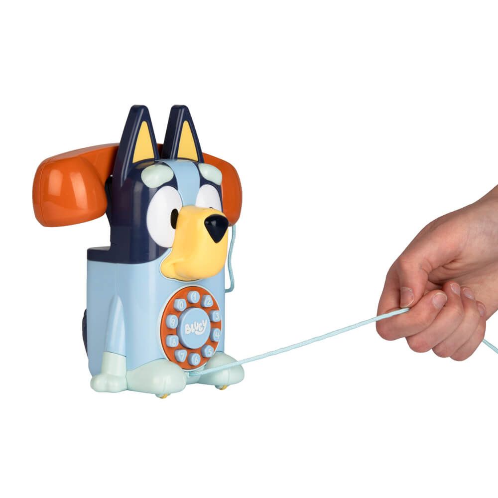 A Bluey-themed toy phone shaped like a dog, with a human hand pulling the cord on the right, set against a plain white background.