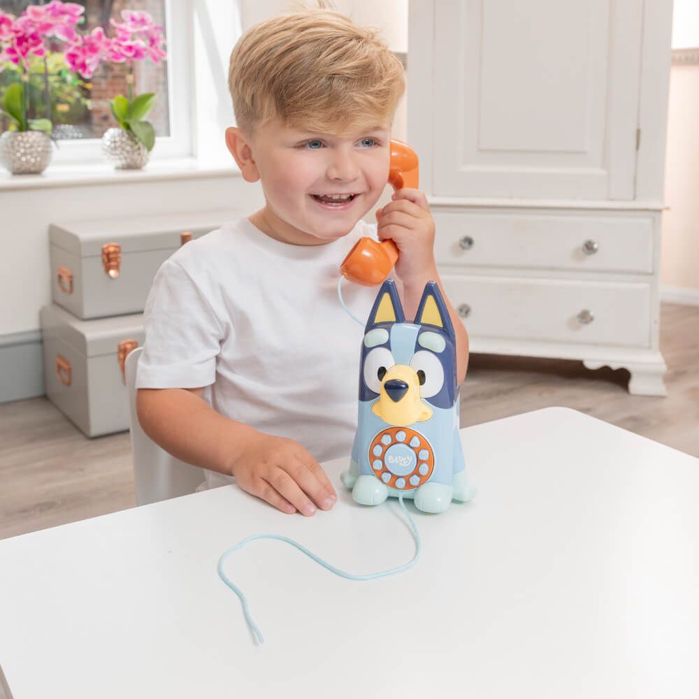 A young boy with blond hair smiling while holding an orange toy phone receiver to his ear. The toy phone is shaped like a blue dog and sits on a white table.
