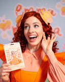 Emma Memma with bright red curly hair wearing a peach collared shirt and orange dress, holding a package of Melissa & Douglass 'In the Barnyard' dog in ears stickers. She is wearing butterfly headbands and smiling excitedly against a light blue background with 'Emma Memma' written on it.