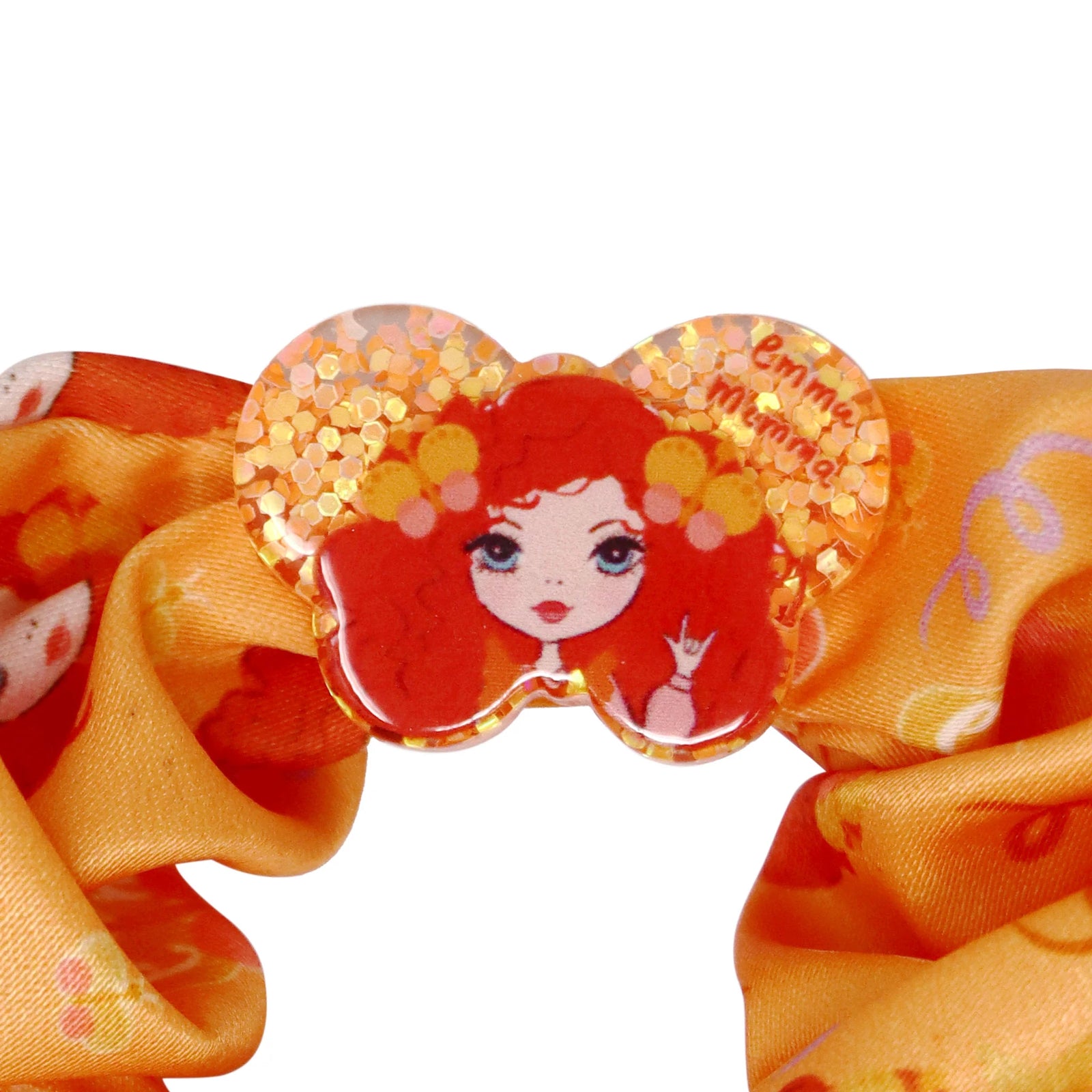 An orange scrunchie with Emma Memma character patterns and a label attached.