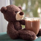 A brown plush teddy bear sitting next to a glass of iced coffee in front of a softly blurred window background.