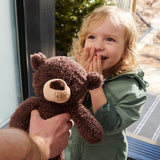 A young child in a green jacket holds a brown plush teddy bear with a beige snout, while another hand supports the bear. The child is smiling and covering their mouth, with an outdoor background visible behind them.