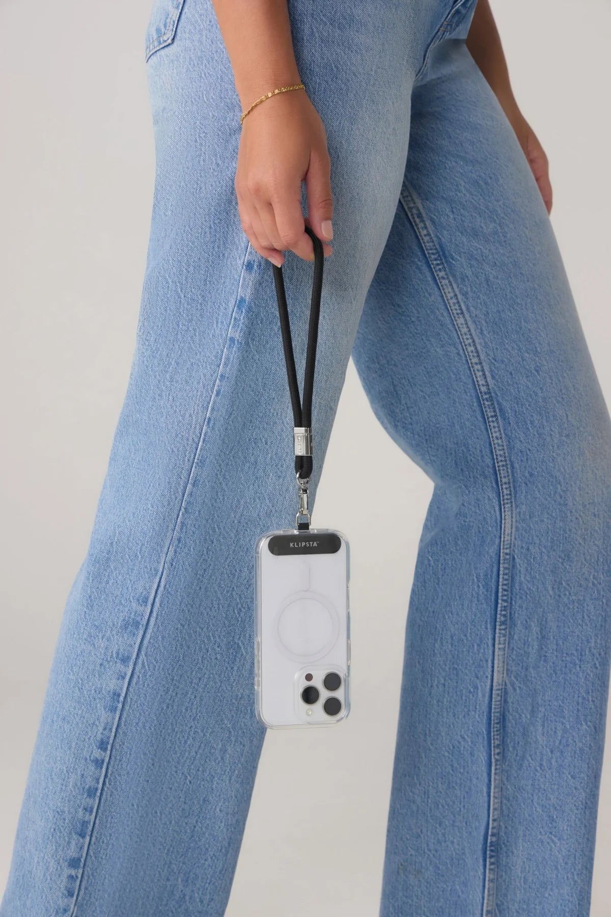 A close-up of a person wearing light blue jeans and holding a black wrist strap attached to a clear phone case with a white MagSafe ring, shown against a plain light background.