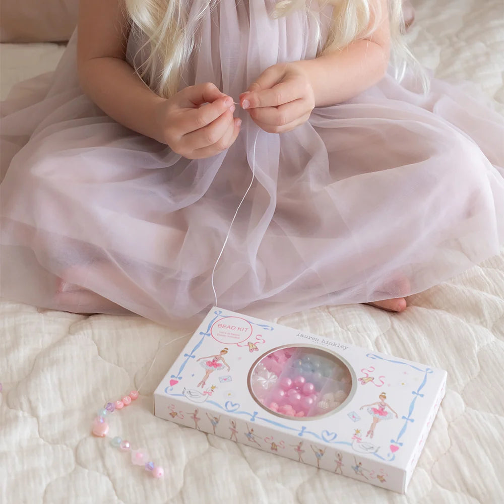 Child in a pink dress sitting on a bed with a box of jewelry and beads.