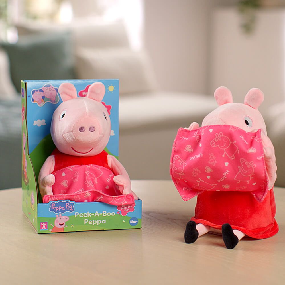 Peppa Pig plush toy shown next to its retail box on a tabletop in a softly blurred indoor setting. The pink pig wears a red dress and holds a pink patterned peek-a-boo cloth, with the box labeled Peppa Pig Peek-A-Boo Peppa.