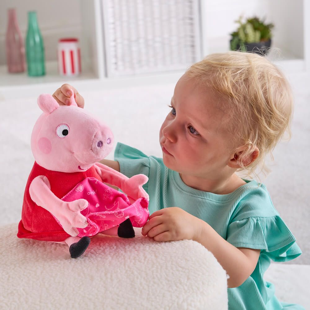 A young child with blonde hair wearing a mint green shirt is interacting with a Peppa Pig plush toy on a white textured cushion in a bright, modern room.