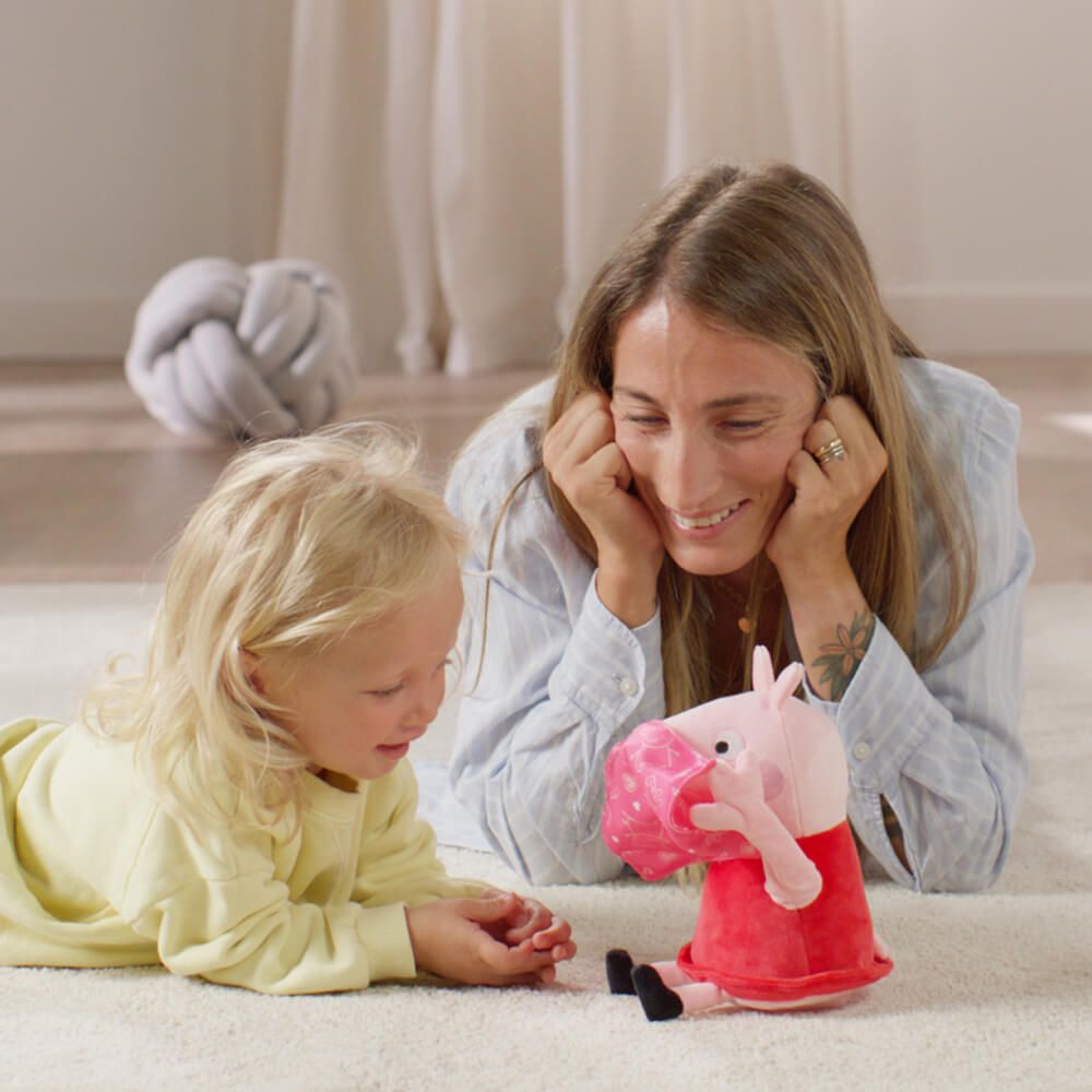 A woman and a young child lying on a carpeted floor, looking at and interacting with a Peppa Pig plush toy in a brightly lit indoor room.
