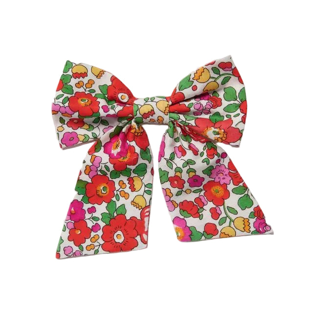 A fabric hair bow featuring a colorful retro floral pattern with red, pink, yellow, and green flowers on a white background, photographed against a neutral light gray backdrop.