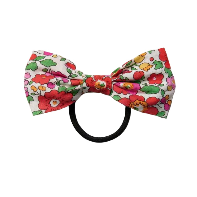 A small bow tie hair accessory with a vibrant red, pink, and green floral print on a white background, attached to a black elastic band, shown against a plain light gray background.