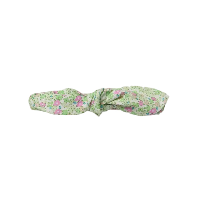 A fabric headband with a knotted bow design, featuring a delicate pink and green floral pattern on a white background, shown against a neutral light gray backdrop.