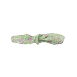 A fabric headband with a knotted bow design, featuring a delicate pink and green floral pattern on a white background, shown against a neutral light gray backdrop.