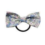 A small bow tie hair clip with a delicate blue, pink, and green floral pattern on a white background, attached to a black elastic band, shown on a light gray background.
