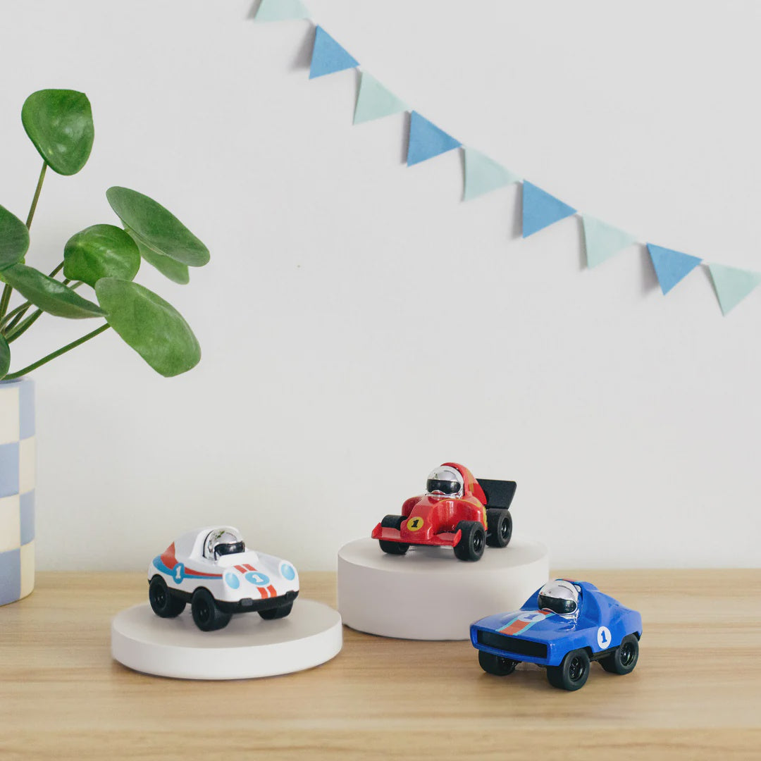 Three diecast toy racing cars in white, red, and blue, displayed on a wooden surface with decorative blue and white triangular garlands in the background.