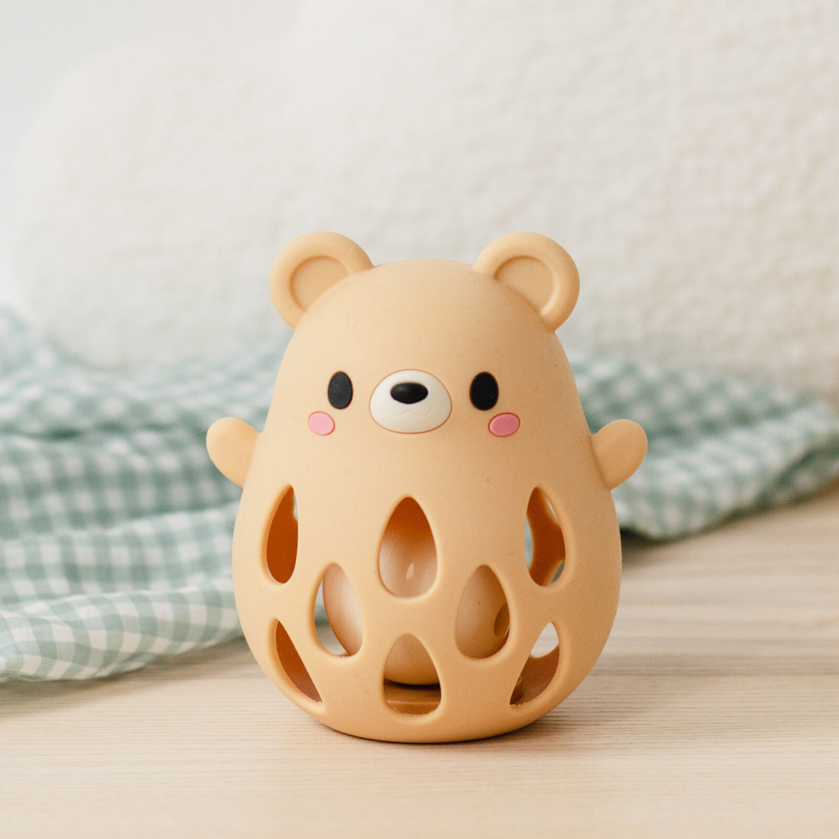 A cute, bear-shaped silicone diffuser or candle holder made of warm beige silicone, featuring a cute cartoon face and teardrop-shaped cutouts on the front. It is styled on a light wooden surface with a green and white checkered cloth and a soft white textured background.