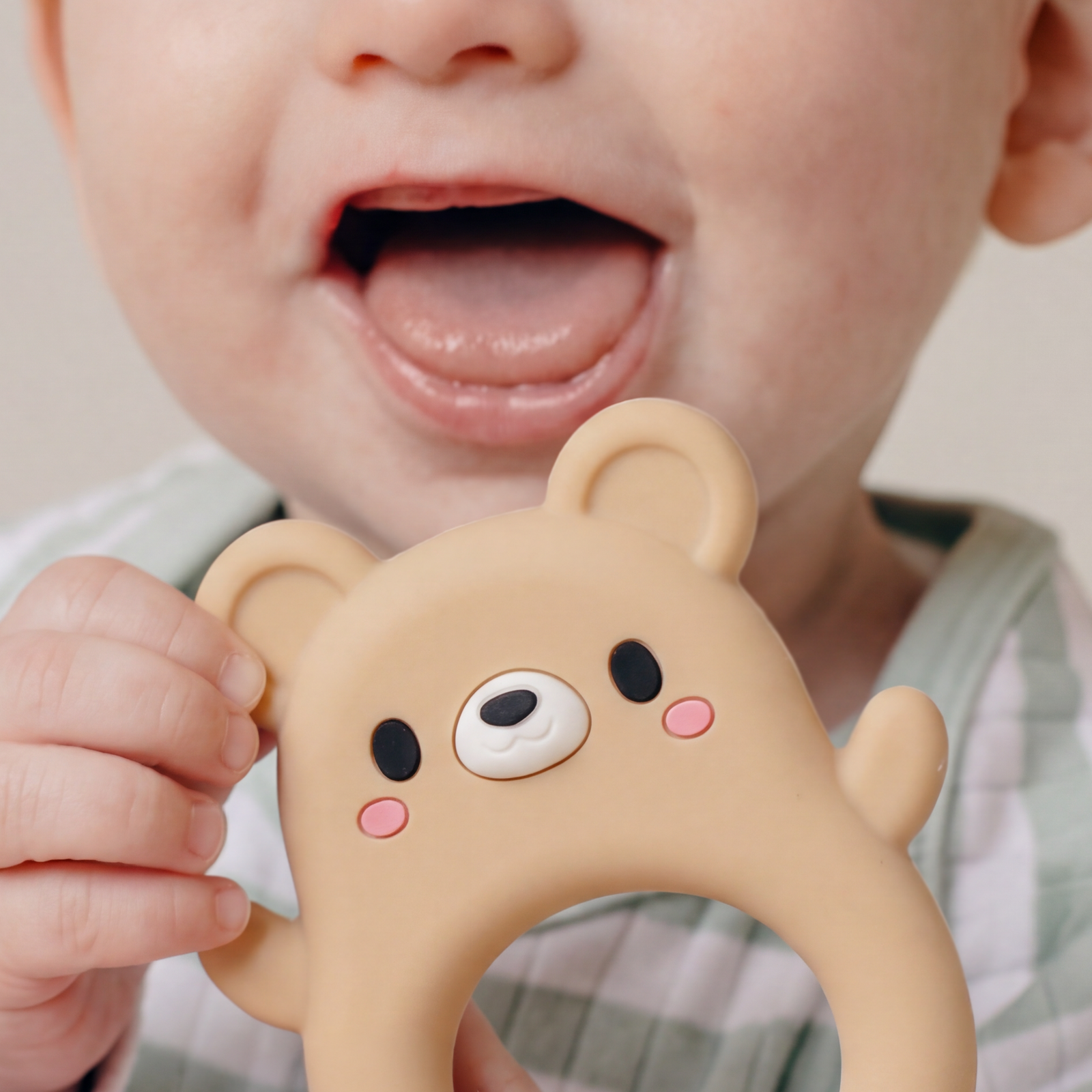 A beige silicone baby teether shaped like a bear face with a circular ring handle, featuring black eyes, a white snout, and pink cheeks, shown on a light gray background.