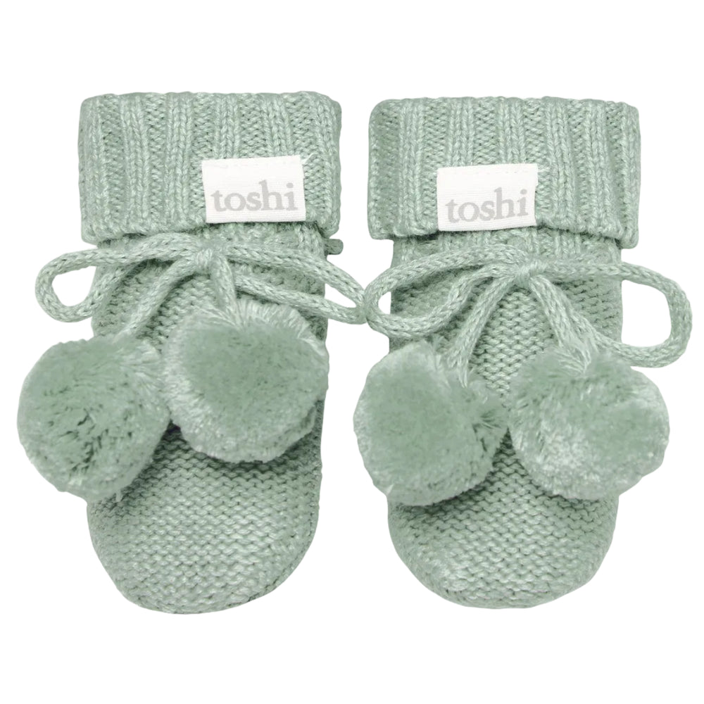 Pair of green baby booties with pom-poms on a white background, featuring the brand 'toshi'.