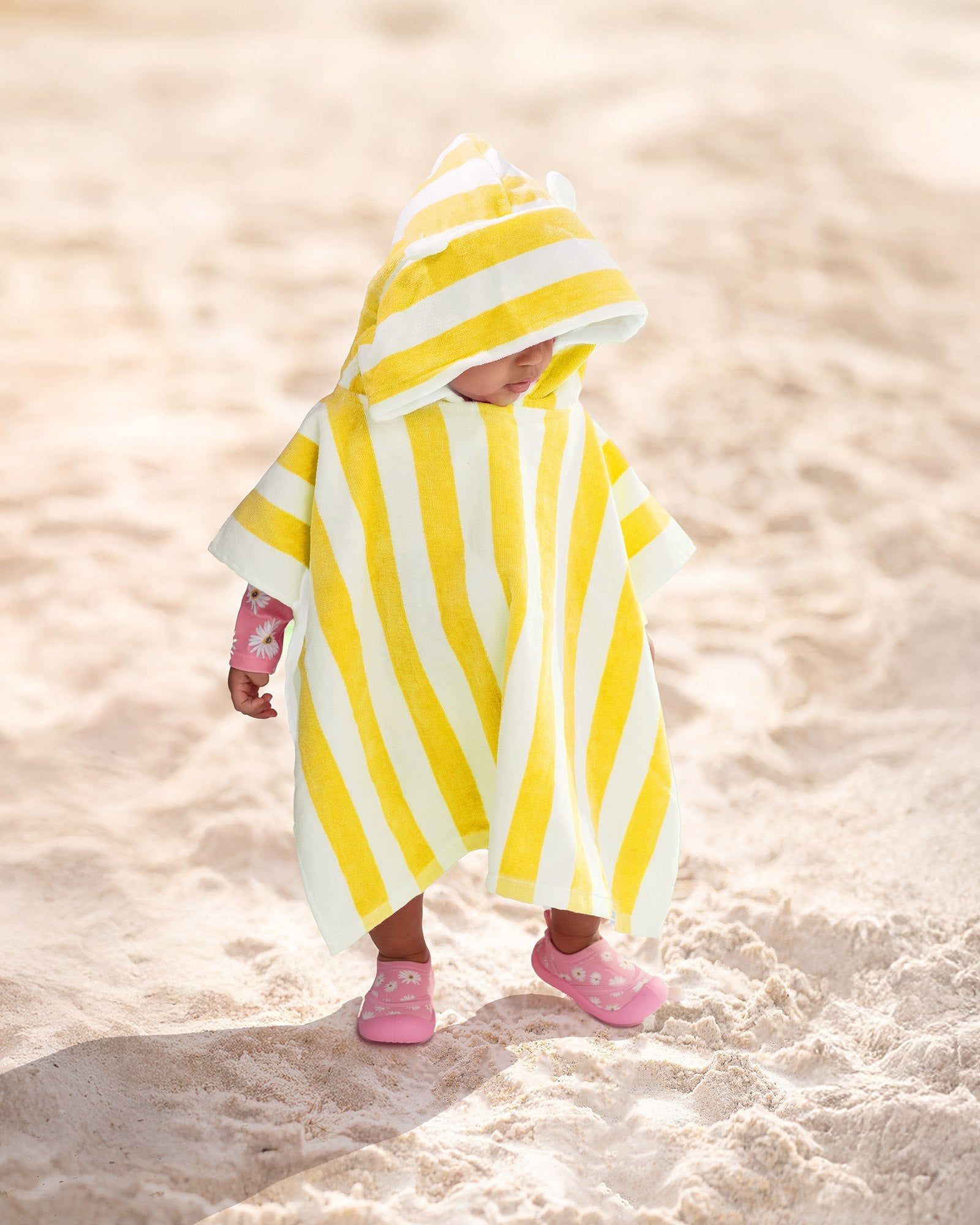 A yellow and white vertically striped beach towel with double shoulder straps and small pom-pom details at the top, shown flat against a light gray background.