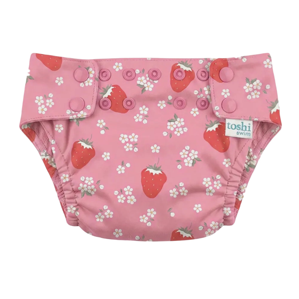 A pink reusable cloth swim nappy with a strawberry and white flower print, featuring three snap closures on the front and a visible 'toshi' brand tag.