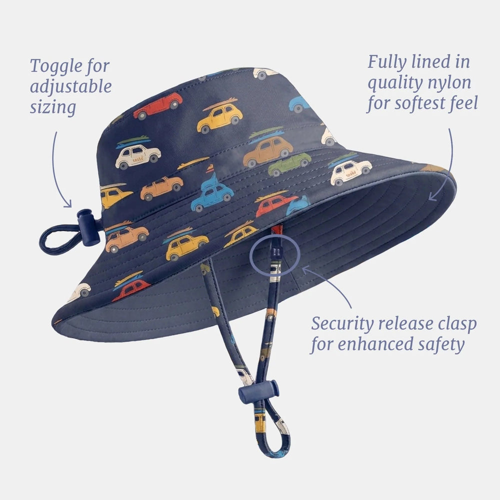 A navy blue swim hat with a colorful vintage car pattern, shown with callout lines and text explaining features like adjustable sizing, fully lined nylon, and a security release clasp, on a light gray background.