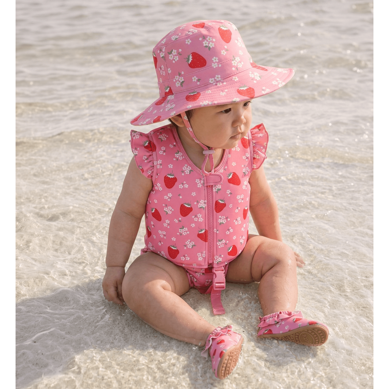 A baby sitting in shallow water on a beach, wearing a pink strawberry and white flower print sun hat, a matching pink ruffle-shoulder one-piece swimsuit with a front zipper, and pink slip-on  beach shoes.