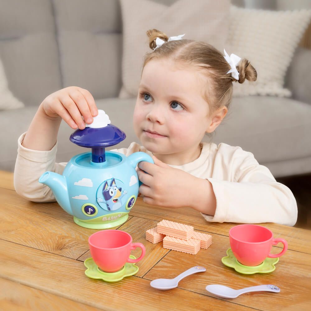 A young child with twin buns and white hair bows is playing with a blue and purple toy teapot and matching pink cups on a wooden table. The toy set features a light blue teapot with a cartoon dog graphic, green saucers, and pink cups, alongside small toy waffles and spoons.