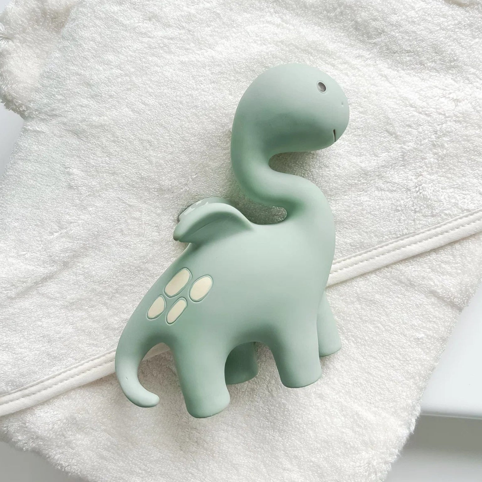 A pale mint green dinosaur-shaped bath and teething toy with small cream spots rests on top of a folded white towel with bear ears.