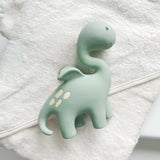 A pale mint green dinosaur-shaped bath and teething toy with small cream spots rests on top of a folded white towel with bear ears.
